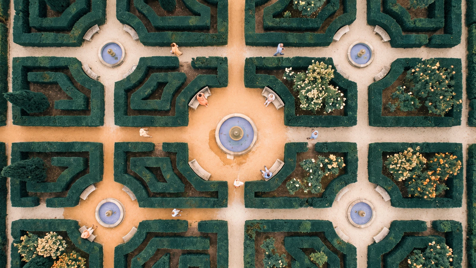 An overhead photograph of a castle garden with a symmetrical hedge labyrinth, five round blue fountains, sandy paths, benches, and walkers.