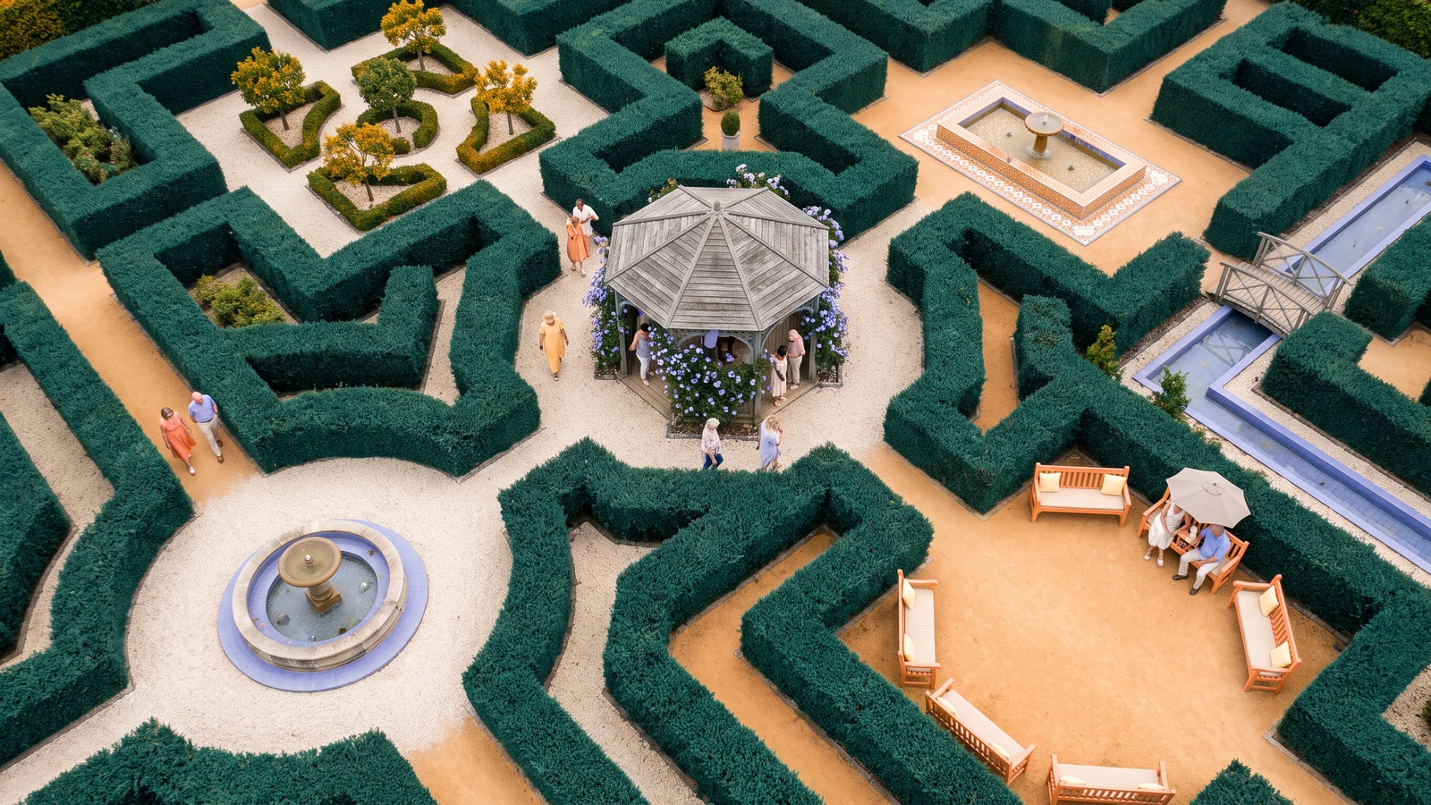 An aerial view of a symmetrical hedge maze with a wooden gazebo in the center, ornamental fountains, garden benches, and visitors walking along sandy paths.