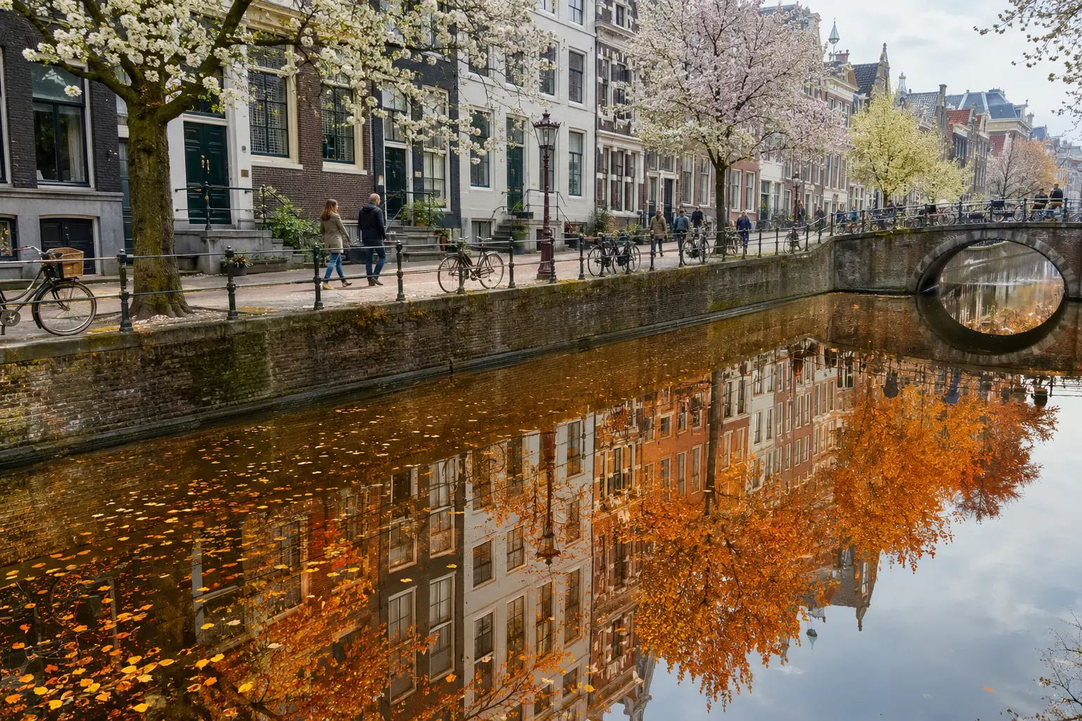 Spring canal with blossoming trees, historic facades, cyclists and a stone arch bridge, while the reflection in the water shows the same cityscape in autumn colours.