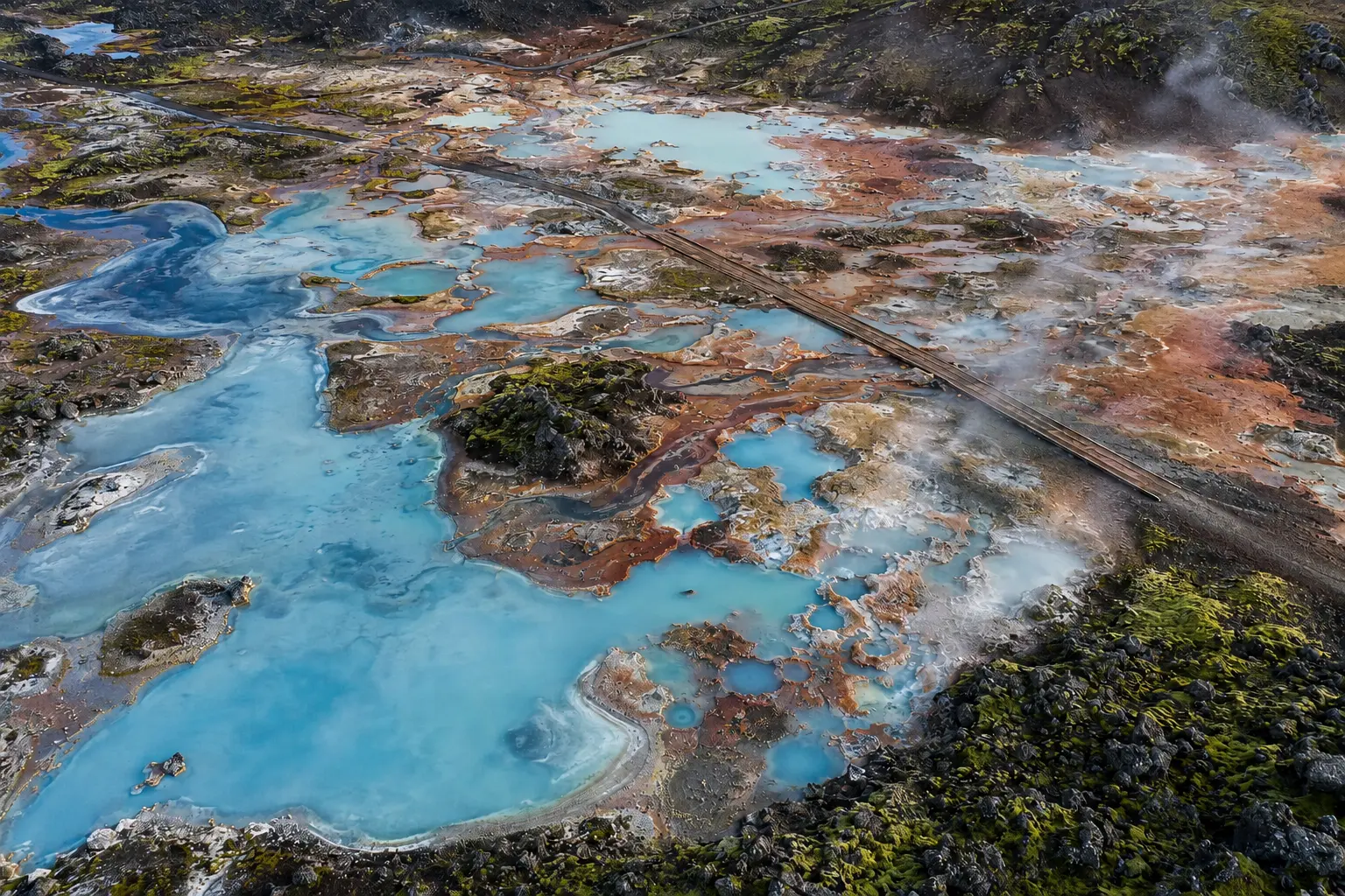 Aerial view of geothermal terraces with clear blue mineral pools, rust-coloured deposits, black lava rocks, green moss fields, steam plumes and a narrow wooden walkway.