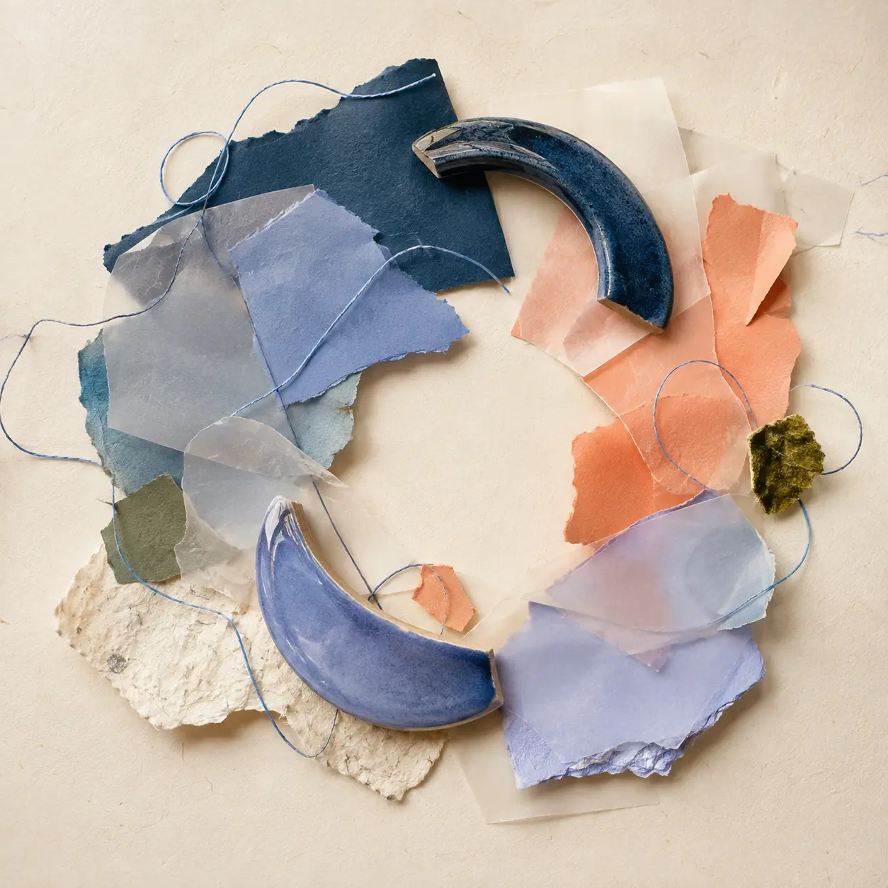 Round studio composition of handmade paper, periwinkle glaze, translucent vellum and loose blue thread on a cream table