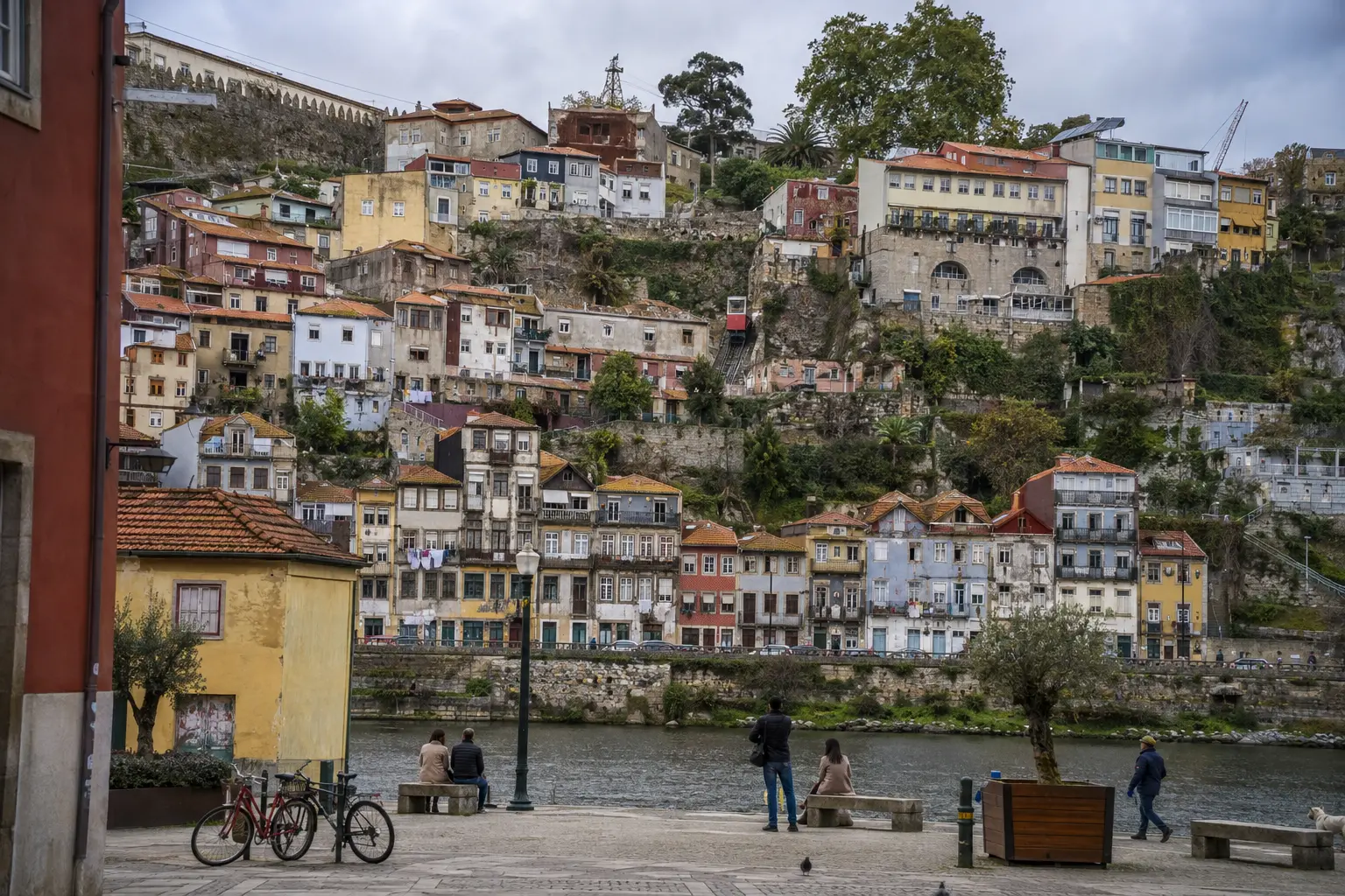 Weathered colourful houses climbing a steep riverbank with a red funicular, bicycles and people along the quay
